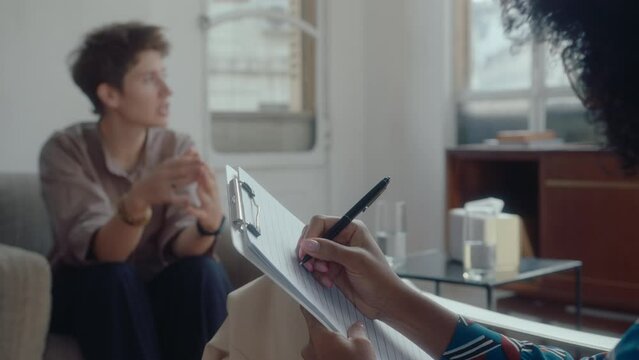 Female psychologist listening to complaints of patient, writing down notes on clipboard during therapy session at clinic. Selective focus, close-up shot
