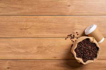 A coffee cup takeaway and many coffee beans are placed around on a wooden table in a warm, light atmosphere, on a dark background, with copy space.