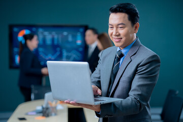Handsome business man and suit holding the laptop in his hands and writing something. Side view, standing confident in the office in front of his team.