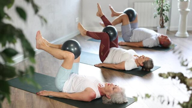 In studio, people of different ages engaged in Pilates. Young girl coach performs exercise with elderly people. Visitior yoga class lying on floor, with bent legs raised and holding softball in knee