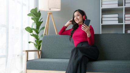 Cute Asian young woman wearing headphones sit on sofa and using smartphone. Asian woman red shirt is using mobile phone, watching movie, listening to music, shopping online, watching live broadcast.