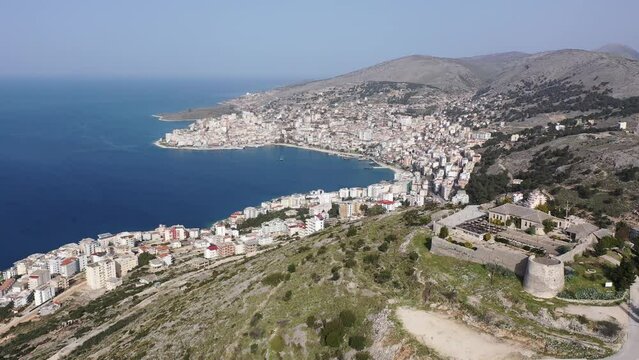Scenic aerial view of Sarande cityscape on shores of gulf of Ionian Sea and ancient Lekuresi Castle on hill above city on sunny spring day, Albania