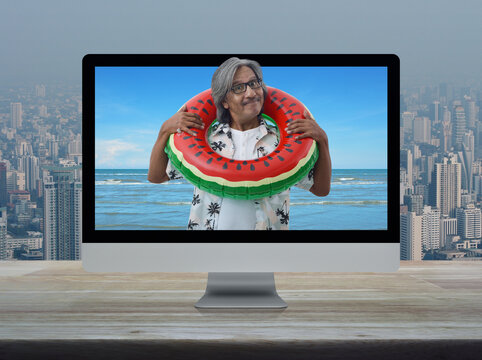 Happy senior traveler asian man wearing red stripped shirt and sunglasses in watermelon inflatable ring with tropical sea on computer screen on table over city tower, Business summer holiday online