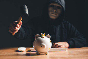 Black hat hacker in mask and hoodie trying to destroy financial piggy bank from digital bank...