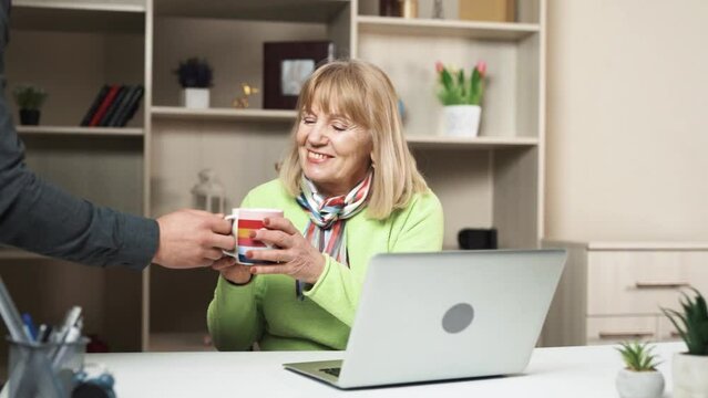 The woman is sitting at the table with a laptop in front of her, working on it. As someone approaches her, they bring a cup of tea or coffee. She looks happy and surprised.