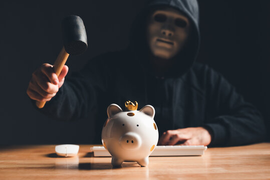 Black hat hacker in mask and hoodie trying to destroy financial piggy bank from digital bank customer dark background. Cyber security and Cybercrime concept. Business investment embezzlement theme.