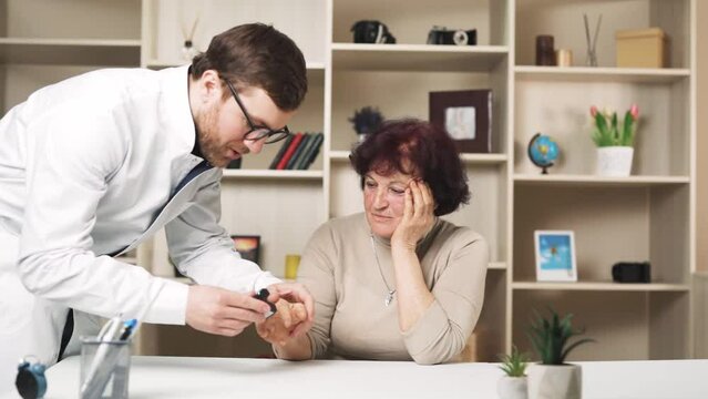 A woman is sitting at a table in a room, feeling unwell. A doctor approaches her and measures her pulse using a stethoscope. The woman is elderly.