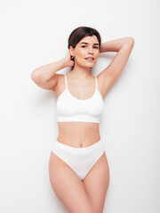 Obraz premium Portrait of young beautiful sexy woman in white lingerie. Smiling carefree model wearing pure underwear. Hot brunette posing near wall in studio interior. Perfect body. Slim and fit