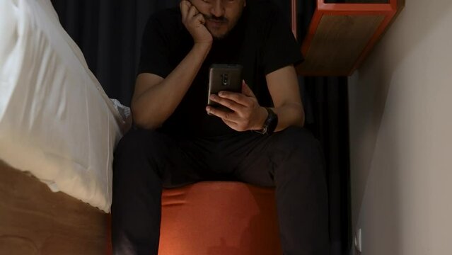 Shot of a man using a mobile phone sitting on a stool beside his bed.