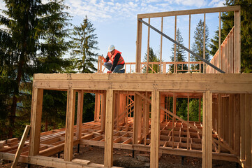 Carpenter constructing two-story wooden frame house near forest. Bearded man hammering nails into...