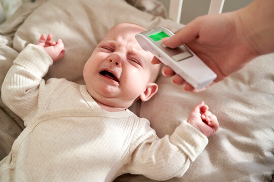 Mother Checking Temperature Of Crying Baby Lying In Crib At Home