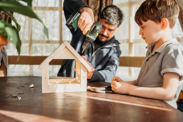 Father drilling birdhouse on table with son at home