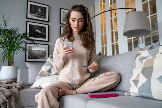 Young woman using smart phone holding pregnancy testing kit at home
