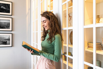 Smiling young woman reading book standing by cabinet at home
