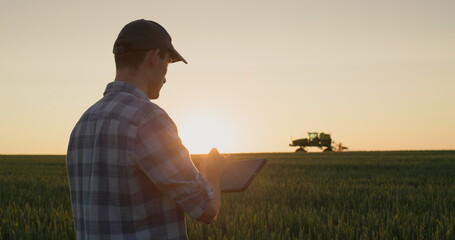 Successful young farmer is working in the field. A tractor is driving in the background. A man uses...