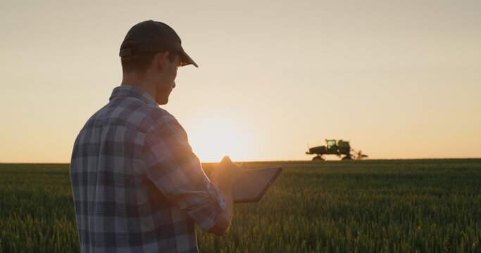 Successful young farmer is working in the field. A tractor is driving in the background. A man uses a tablet - new technologies in agriculture - Powered by Adobe