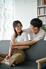 An Asian man embracing his girlfriend from behind while relaxing in the living room together.