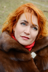 red-haired woman wearing white knitted hat and dressed in warm hooded casual parka jacket outerwear, girl in the parking lot, pedestrian crossing and car in the background, girl warms frozen © Oleksandra