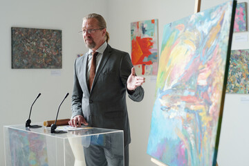 Mature gallery owner in elegant suit presenting new art on canvas in art gallery