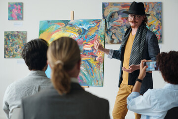 Artist in elegant clothing presenting his picture on canvas to group of people during exhibition in...