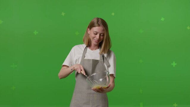 Caucasian woman beats fresh eggs into the glass bowl and puts egg shells to the small dish on green screen background. Female stands in her kitchen