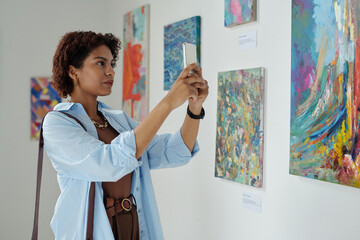 African American young woman making photo of paintings on her smartphone in art gallery