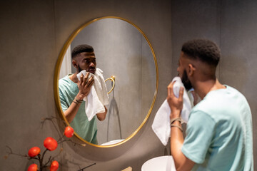 Displeased young African American man wipes face after washing. Male examines derma in mirror after...