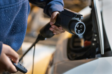 Woman hands holding electric car charger and remote controller, ready to connect to electric vehicle at home garage.