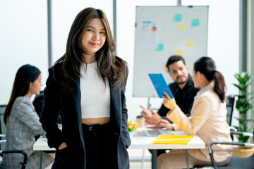 Portrait of Asian business woman stand with relax position and look at camera in front of other co-workers discuss on the back in office.