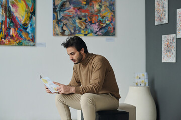 Young man resting on couch and reading brochure at exhibition of modern art in gallery