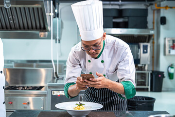 Asian male chef cooks macaroni and decorates the dish beautifully with vegetables to look appetizing before serving to customers using smartphones to take pictures of the food in the restaurant
