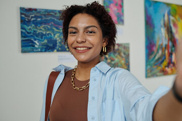 Portrait of African American woman smiling at camera while making selfie portrait against paintings...