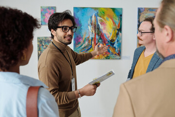 Young guide pointing at paintings on the wall and talking about modern art to people in gallery
