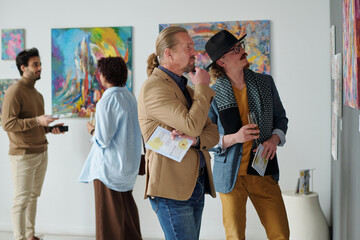 People looking at paintings on the wall and discussing them while visiting exhibition of modern art in gallery