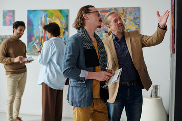 Art lovers discussing modern art in gallery, they pointing at wall with paintings and talking to...