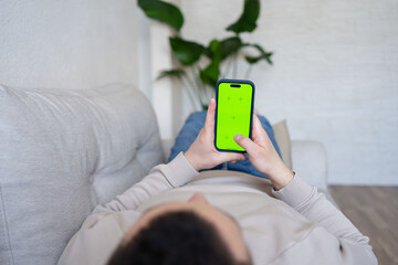 Young man at home resting on a couch using with green mock-up screen smartphone. Guy using chroma key mobile phone, internet browsing, posting on social networks. 