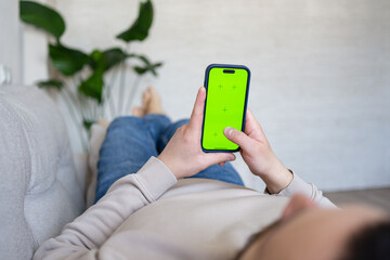 Young man at home resting on a couch using with green mock-up screen smartphone. Guy using chroma key mobile phone, internet browsing, posting on social networks.