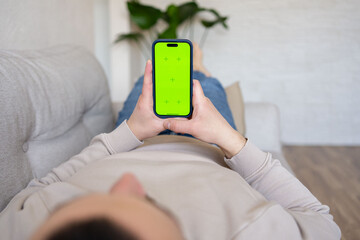 Young man at home resting on a couch using with green mock-up screen smartphone. Guy using chroma key mobile phone, internet browsing, posting on social networks. 