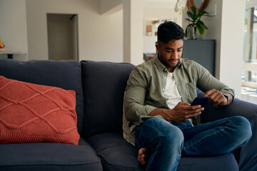 Young biracial man in casual clothing sitting on sofa using mobile phone at home