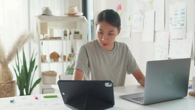 Young Asia girl IT development programmer typing on keyboard coding programming fixing data code on laptop computer screen follow code plan in tablet and paperwork on table workroom at house office.