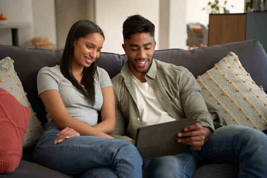 Happy young biracial couple in casual clothing sitting on sofa using digital tablet at home