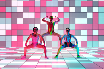 Three men in stylish, vintage sportswear training, doing aerobics exercises over multicolored mosaic studio background in neon light. Concept of sportive and active lifestyle, humor, retro style. Ad