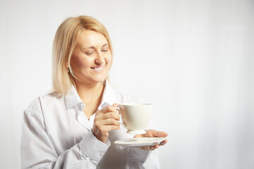 Portrait of pretty blonde smiling woman posing with cup of tea or coffee on white background. Happy girl model in white shirt in studio. The concept of pleasant morning at home or at work. Copy space
