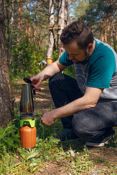 A man uses a travel-sized, compact gas burner to make coffee in a geyser coffee maker. Coniferous forest, rest of the wild nature. Vertical photo