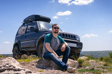 Happy traveler man sitting on rock in sunglasses. An off-road vehicle with a roof rack in the background. The concept of an active lifestyle and recreation. A trip by car to wild places. © Sergey