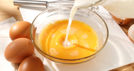 Adding milk to eggs. Milk is poured into a bowl with whipped eggs. Breakfast preparation. Omelet recipe. Rustic style. Sunny day. Close up.