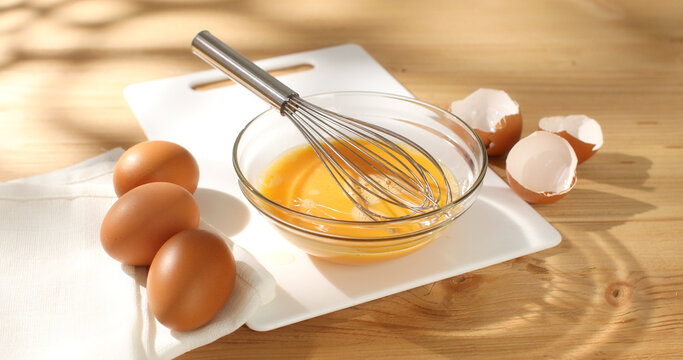 Whisking eggs. Whipped raw eggs in a bowl with whisk. Breakfast preparation. Omelet recipe. Rustic style. Sunny day. Close up.