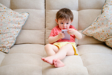 Toddler baby is tapping away on the mobile phone screen. The happy youngster is glued to the screen of their phone while seated comfortably on the couch. Kid aged about two years (one year ten months)
