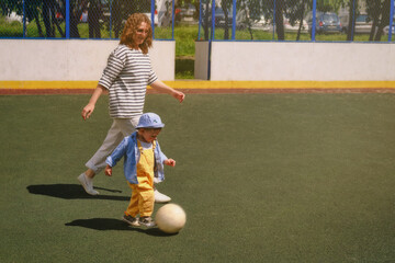 The child and woman mother is having fun kicking the soccer ball around on the outdoor field. Baby with mom is happily playing with the white ball in the park. Kid aged about two years