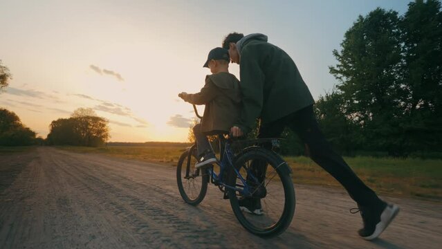 The younger brother learns to ride a bicycle at sunset with the older brother.